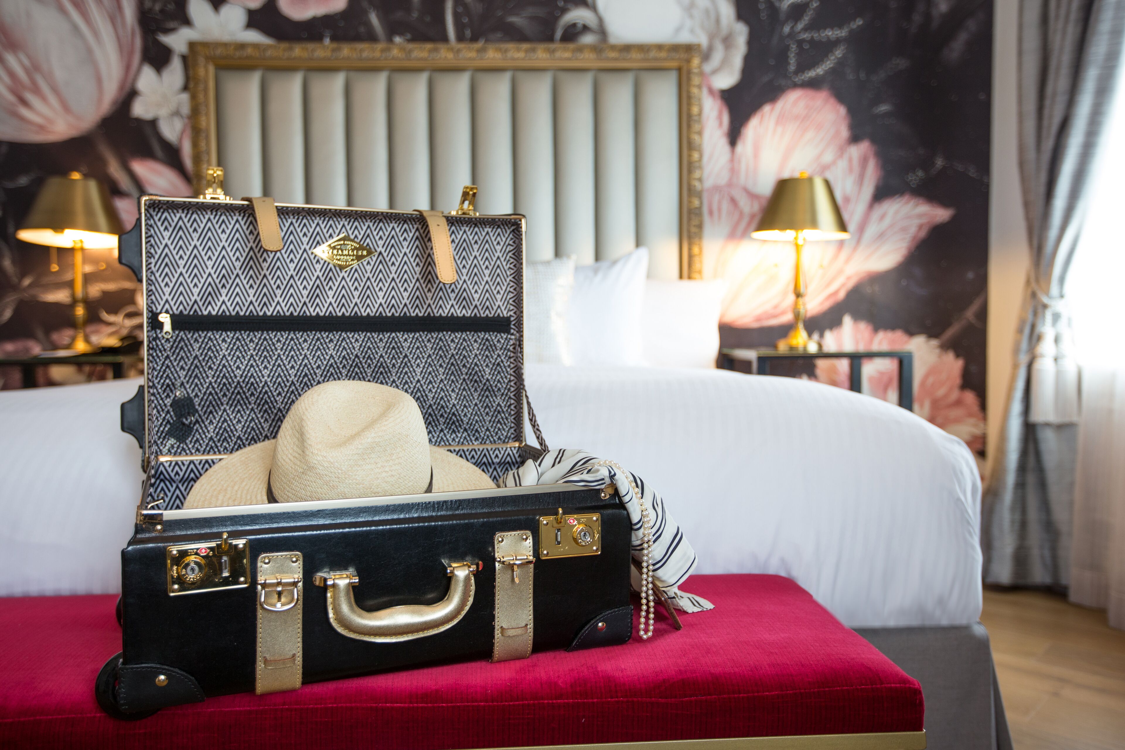 A bed with a red upholstered headboard, a suitcase with a hat, and two bedside lamps.