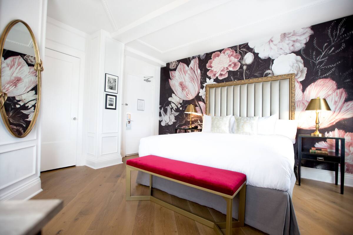 A bedroom with a large bed, a bench, a mirror, and a wall with floral wallpaper.