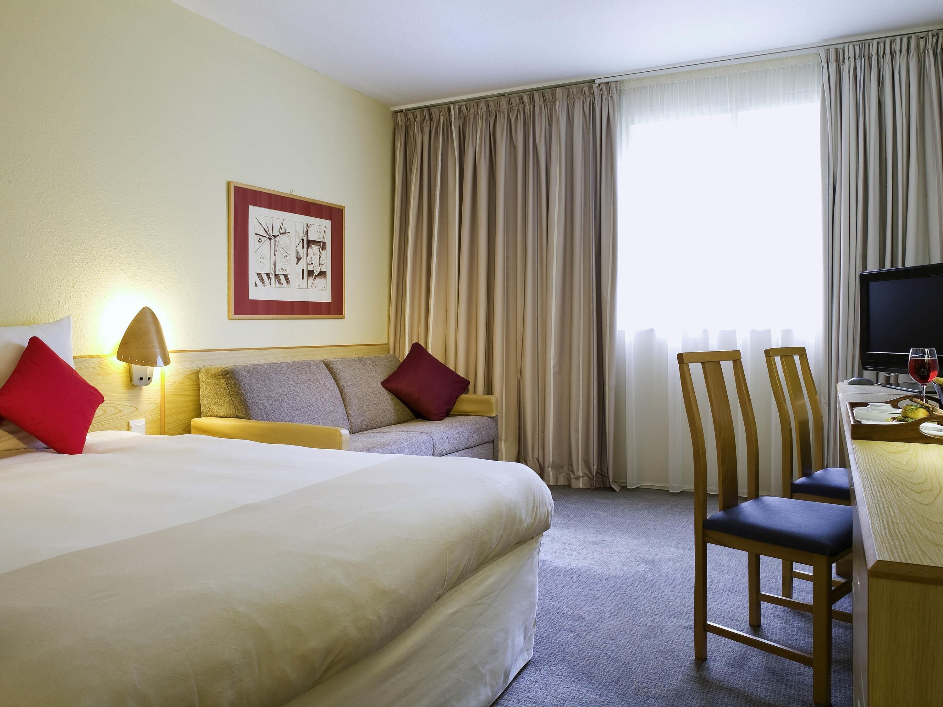 A hotel room with a bed, a sofa, a desk with a chair, a television, and a window with curtains.