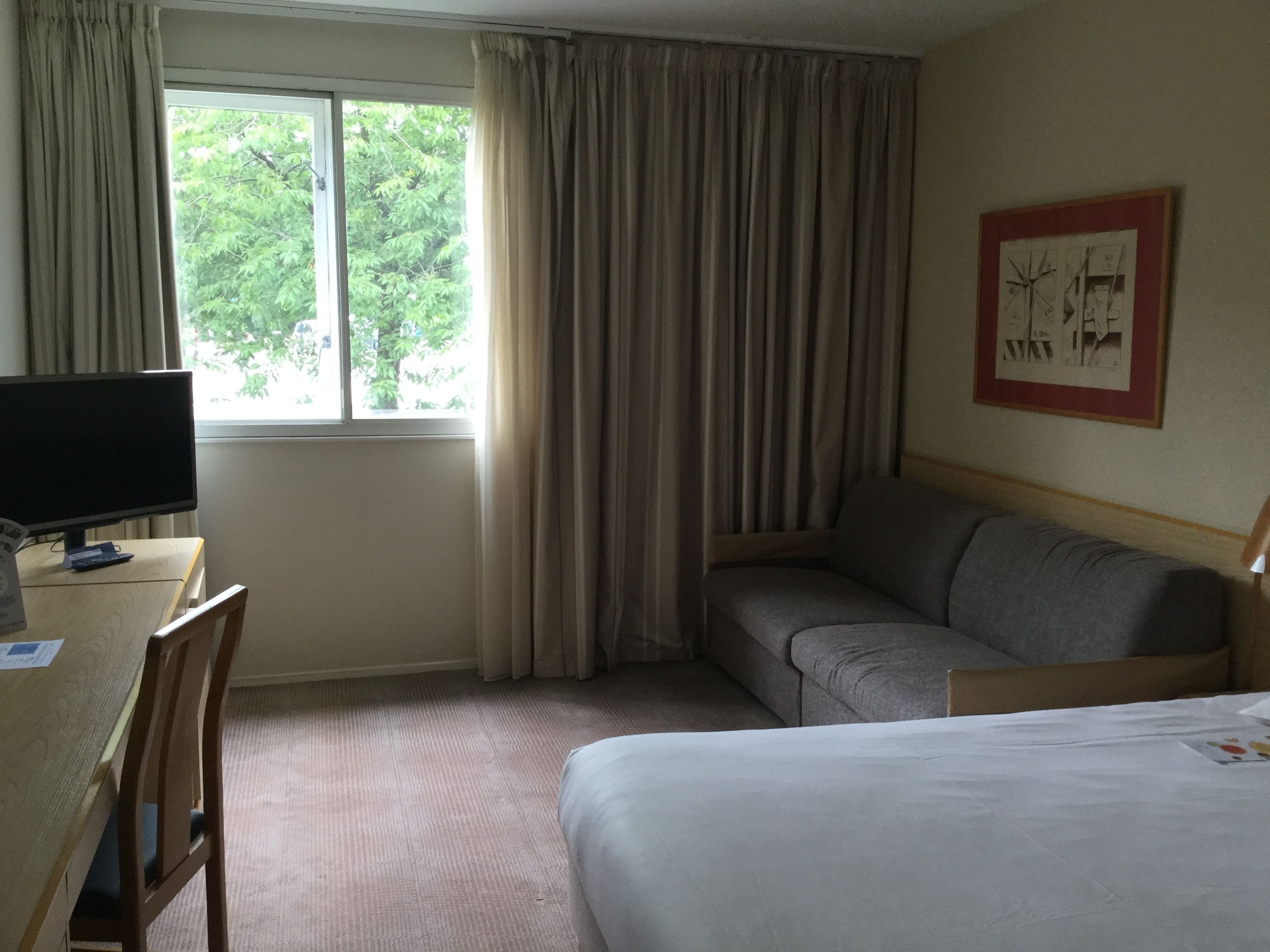 A hotel room with a bed, a desk with a computer, a chair, a sofa, and a window with curtains.