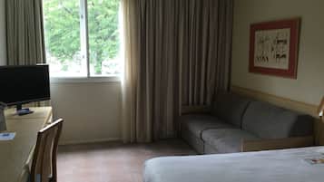 A hotel room with a bed, a desk with a computer, a chair, a sofa, and a window with curtains.