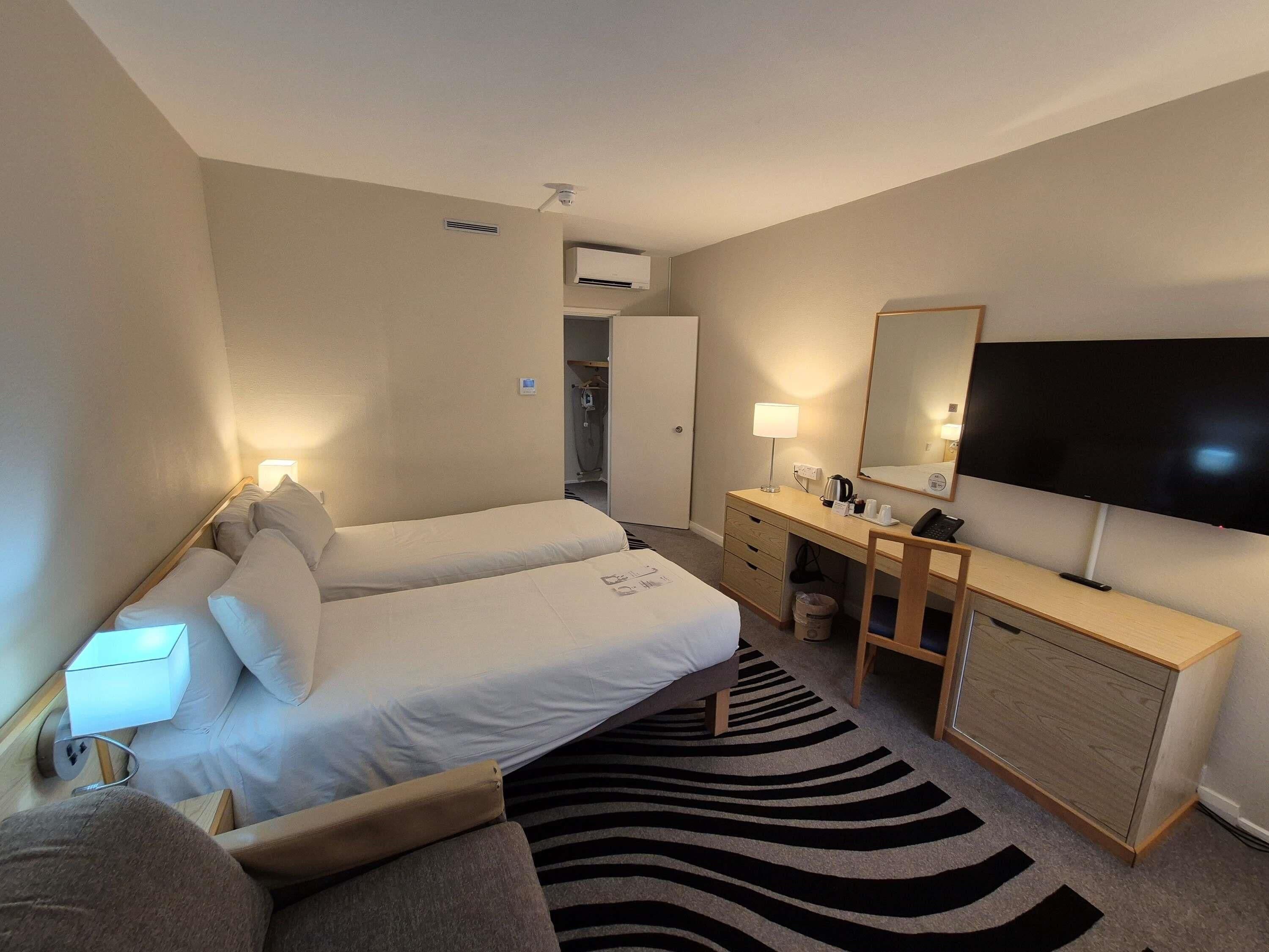 A hotel room with two beds, a desk, a TV, and a bathroom in the background.