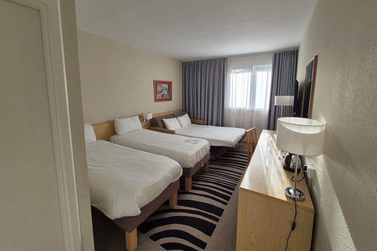 Ein Hotelzimmer mit zwei Betten, einem Schreibtisch mit Lampe, einem Spiegel und einem Fenster mit Vorhängen.