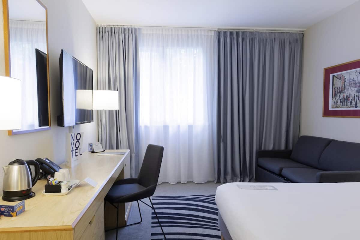 Ein Hotelzimmer mit Bett, Schreibtisch mit Telefon und Wasserkocher, Sessel, Sofa und einem großen Fenster mit Vorhängen.