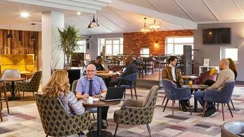 A modern indoor space with round tables and patterned chairs, a brick wall, and a ceiling with exposed beams.