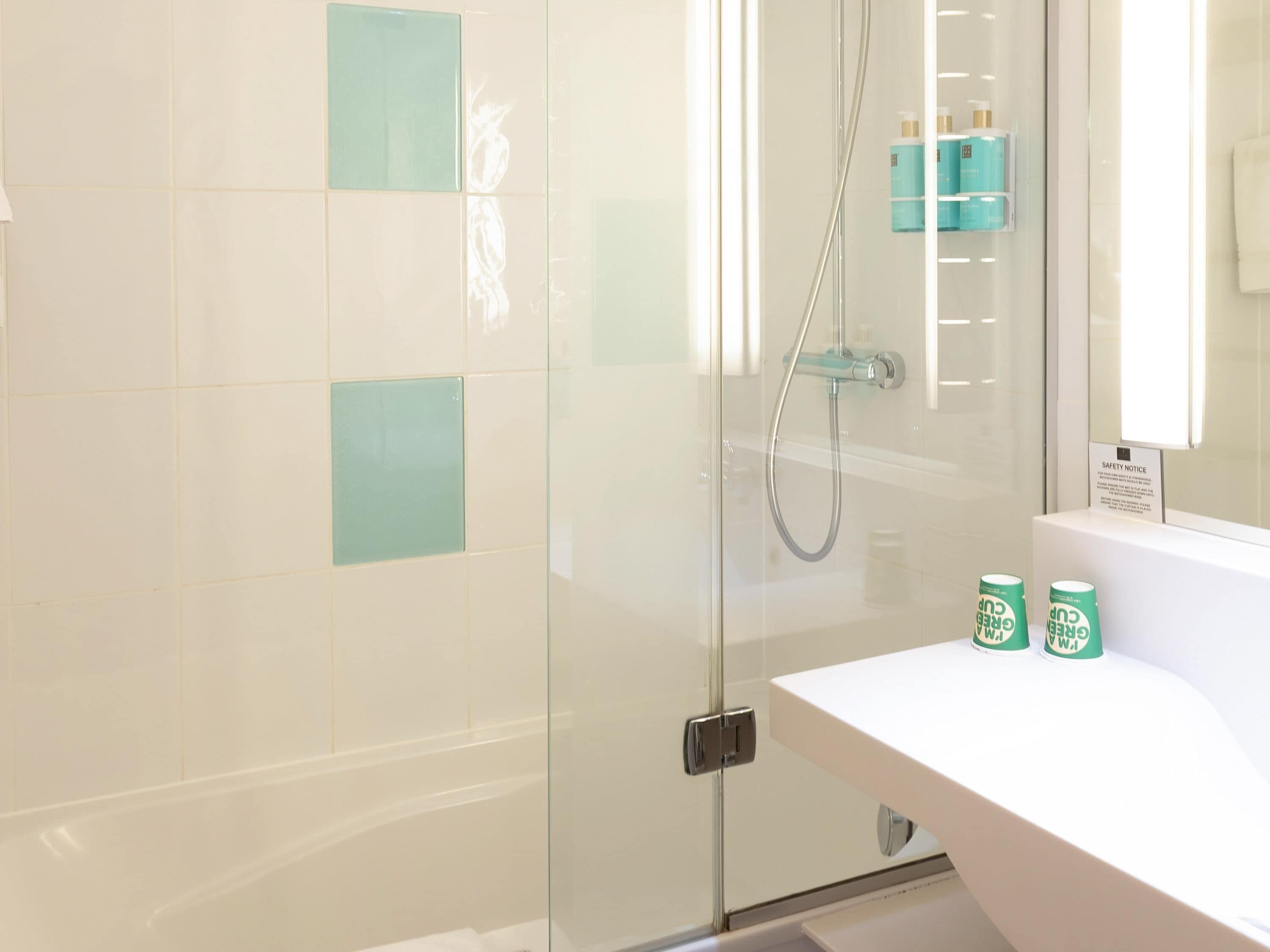 Une salle de bain moderne avec une douche à parois de verre, un lavabo blanc avec un miroir et un panneau d’avertissement de sécurité.