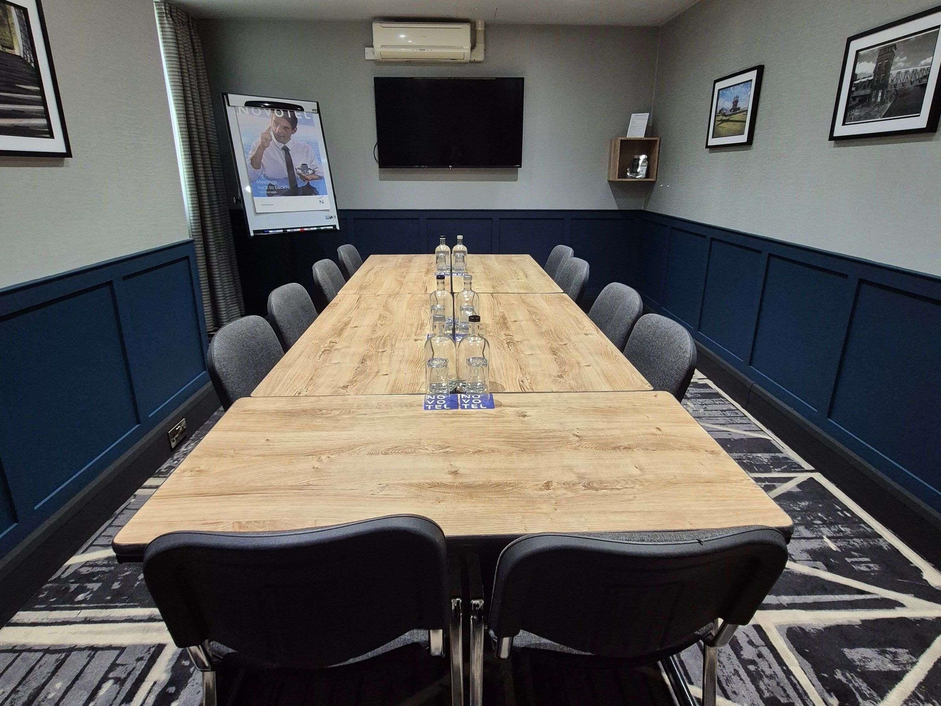 A conference room with a long table, chairs, a projector screen, and framed pictures on the walls.
