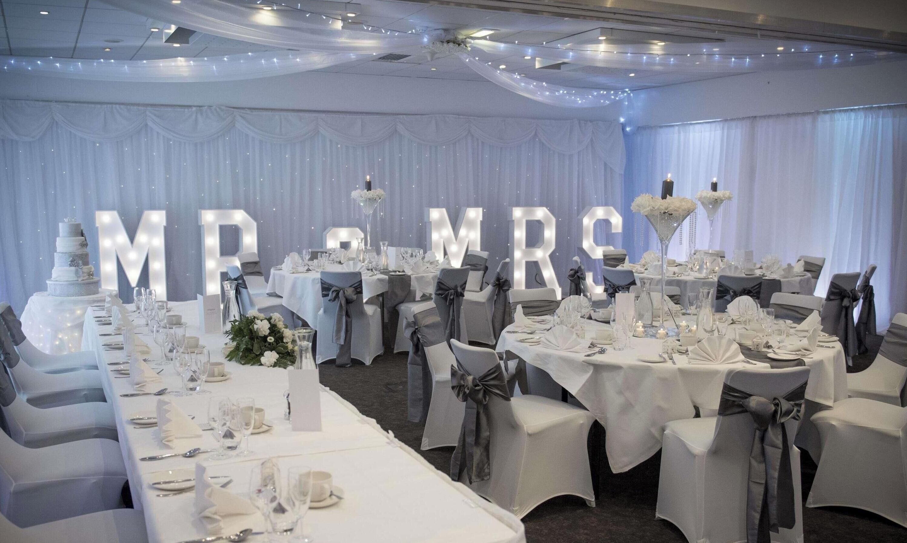 Un salón de banquetes con mesas dispuestas para una recepción de boda, que presenta cortinas blancas, candelabros y letras iluminadas de gran tamaño que deletrean 'M & M R E'.