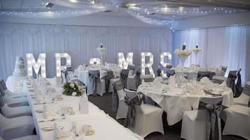 Un salón de banquetes con mesas dispuestas para una recepción de boda, que presenta cortinas blancas, candelabros y letras iluminadas de gran tamaño que deletrean 'M & M R E'.