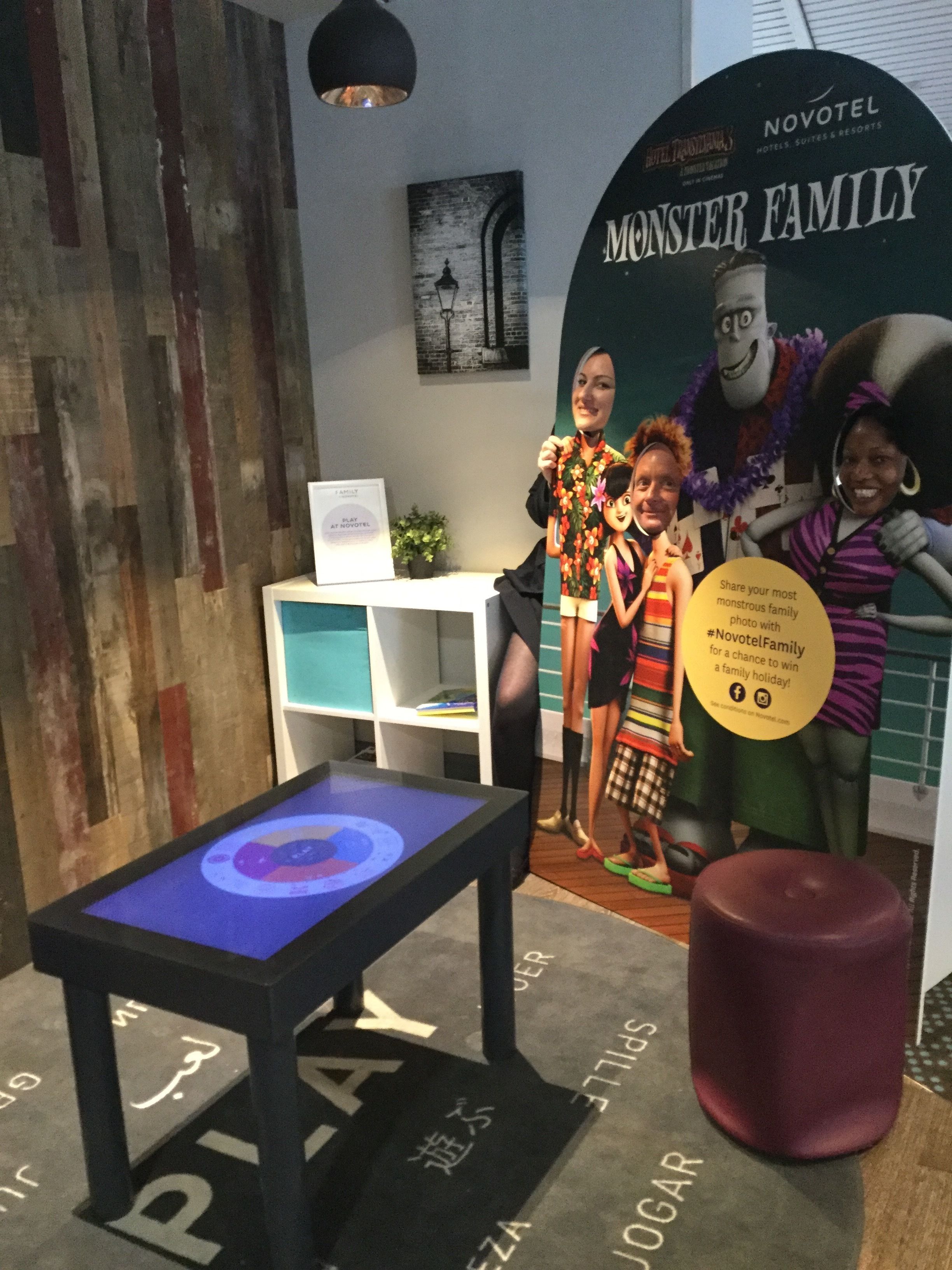 A display area with a Monster Family poster, a table with a circular design, and a sign encouraging visitors to share photos.