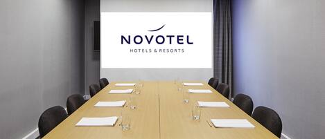 A conference room with a long table, chairs, a projector screen displaying 'NOVOTEL HOTELS & RESORTS', and a carpeted floor.
