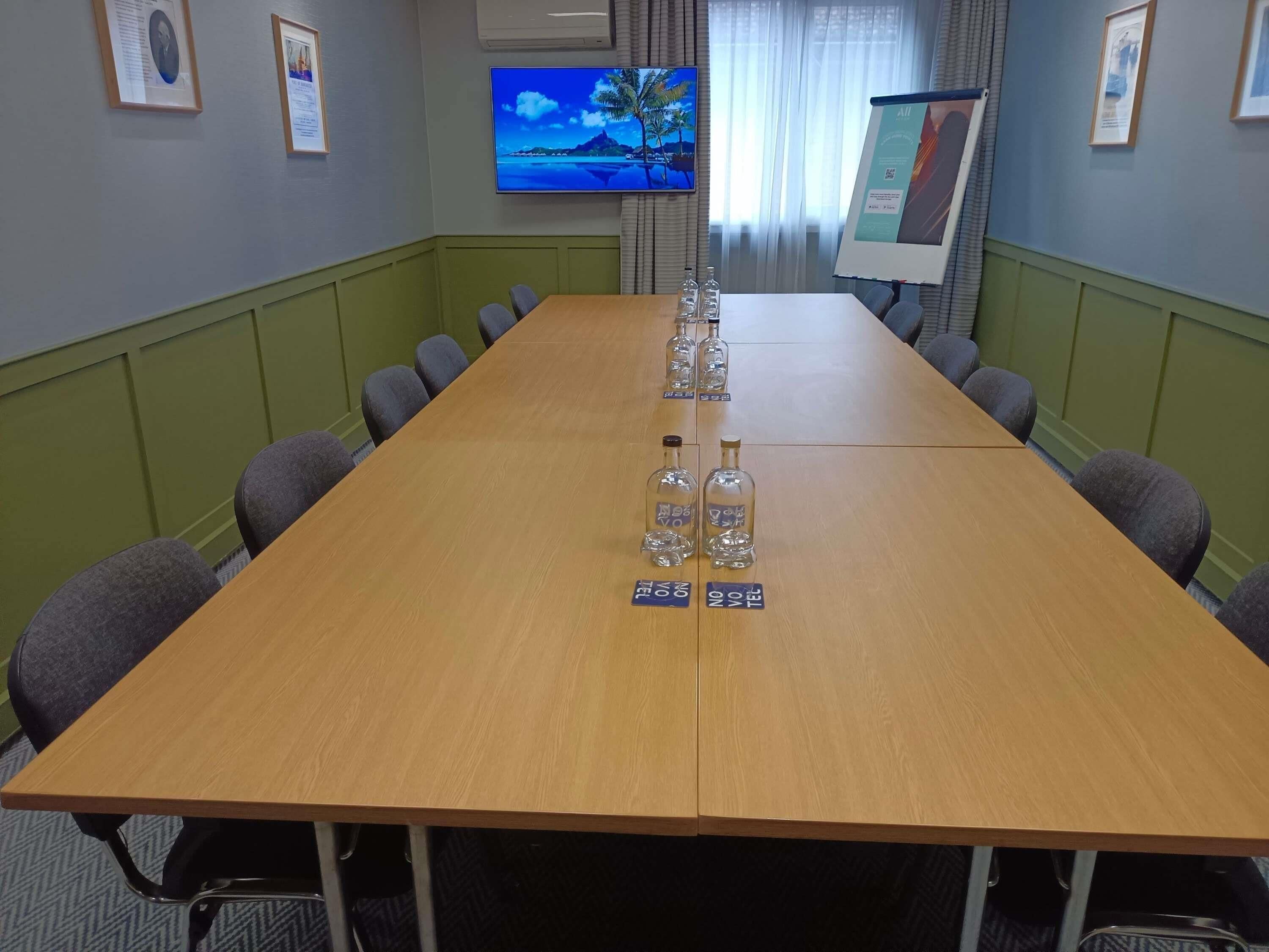 Una sala de conferencias con una larga mesa, sillas, un televisor y una pantalla para presentaciones.