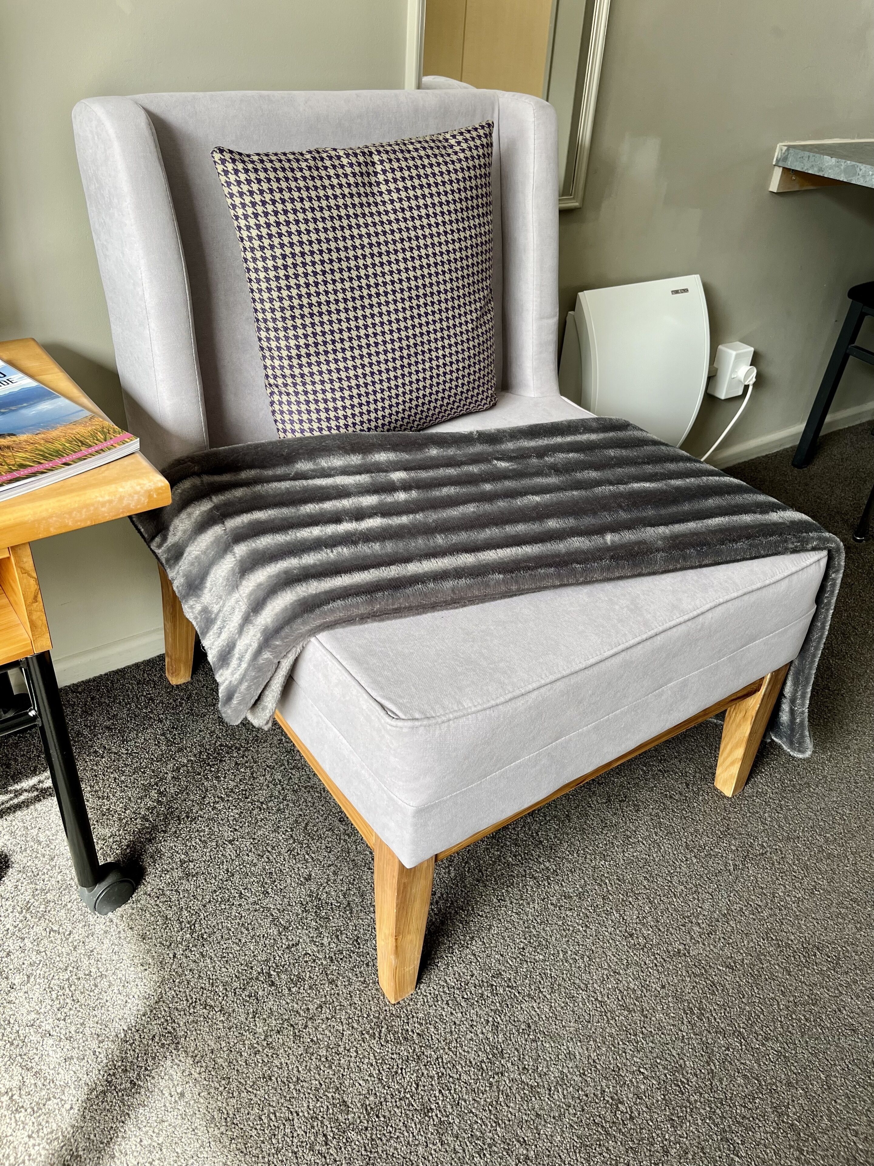 Een grijze fauteuil met een kussen met hondentandpatroon, een grijze deken en een houten bijzettafeltje met een boek.