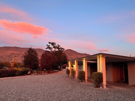 Un edificio con camino de grava y un espacio al aire libre cubierto, rodeado de árboles y montañas al fondo.