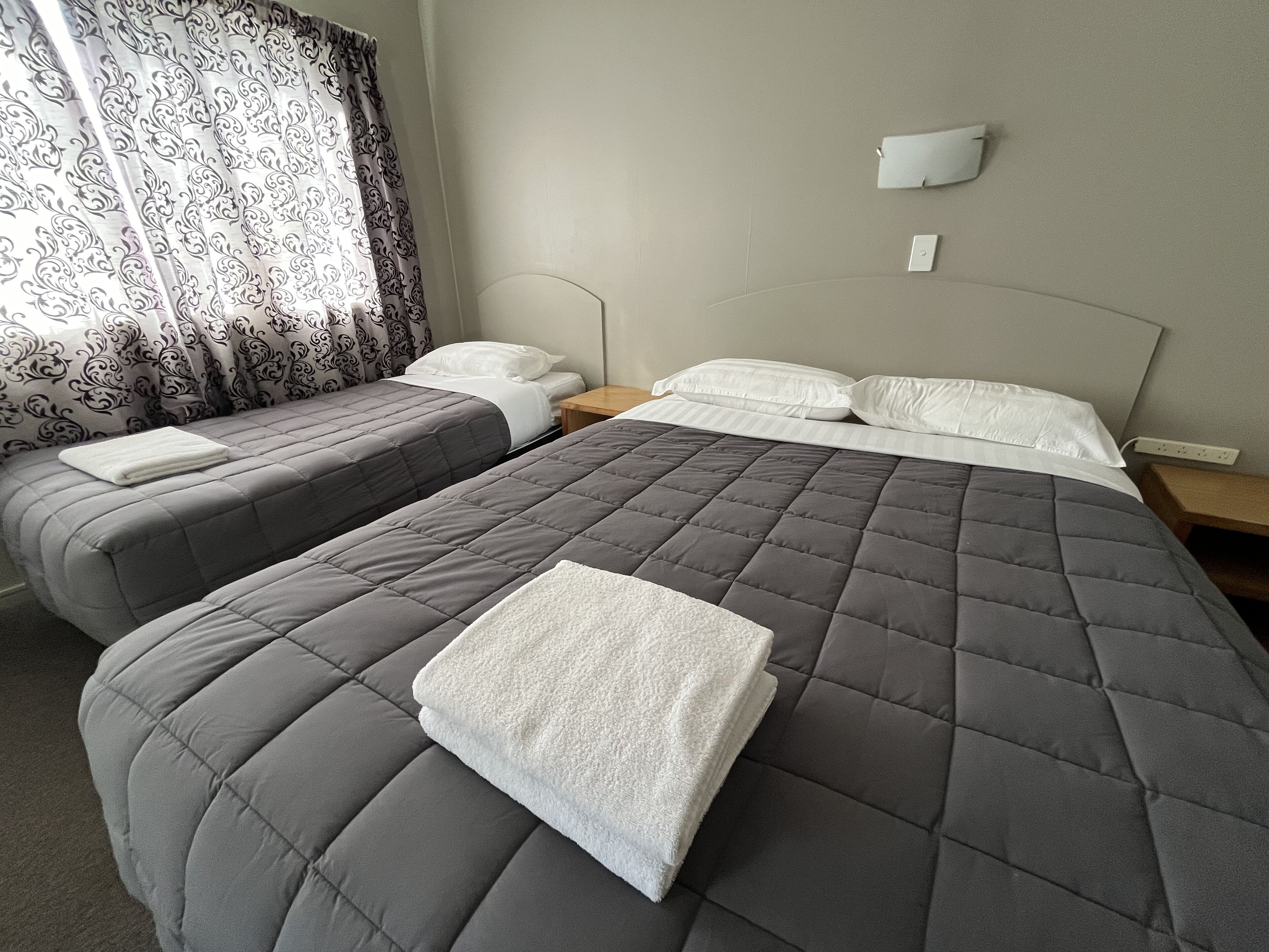 Quarto de hotel com duas camas, cada uma com colcha cinza acolchoada e almofadas brancas. Há uma janela com cortinas estampadas, uma mesa de cabeceira com abajur e um ponto de luz fixado na parede.