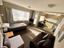Una habitación de hotel compacta con cama, sofá, una mesita, televisión, kitchenette y balcón con cortinas.