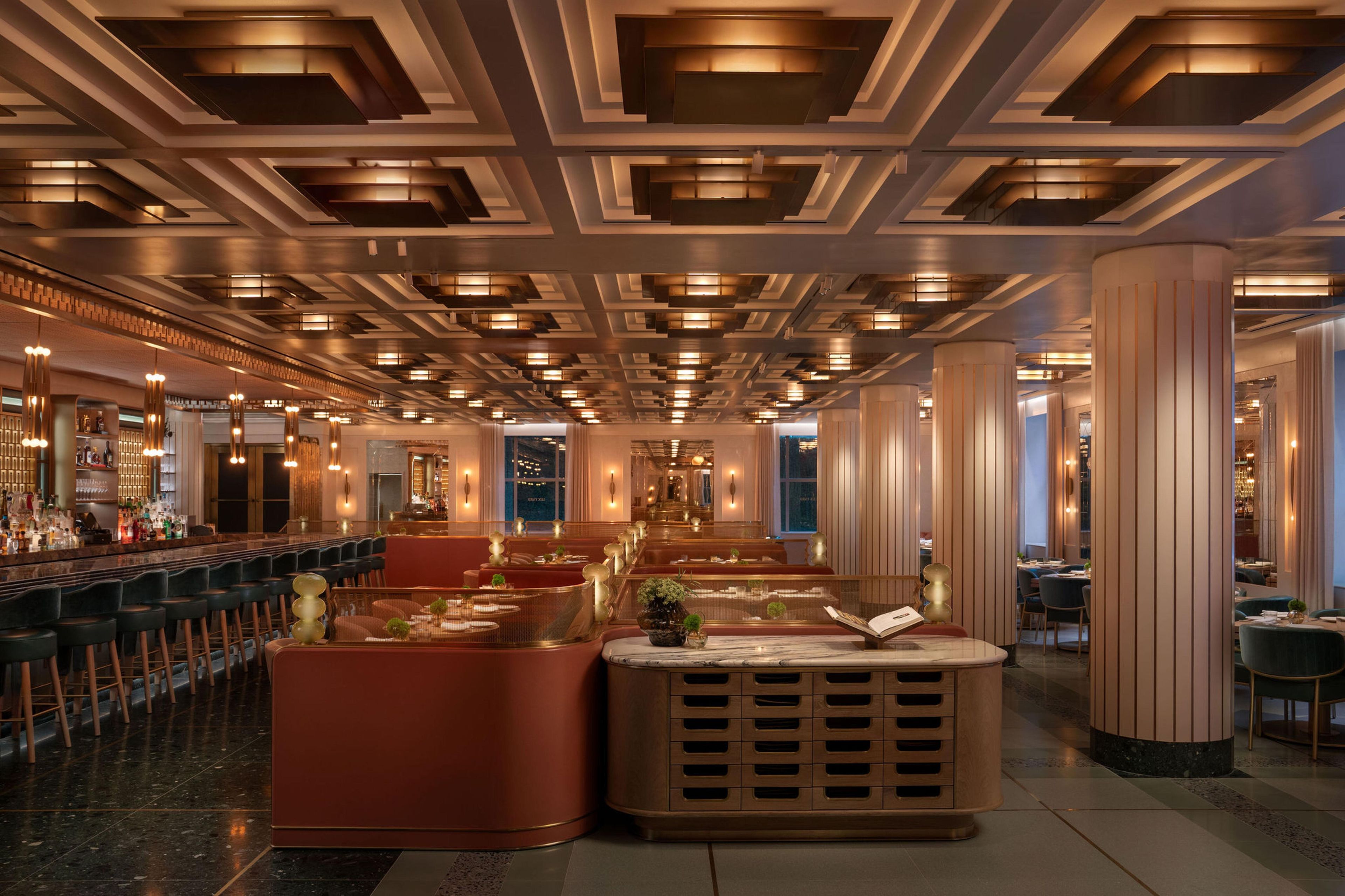 A modern hotel lobby with a long bar, seating areas, and a patterned ceiling.