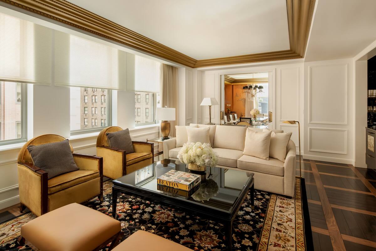 A spacious living room with a beige sofa, two armchairs, a coffee table with books, and a large window with blinds.