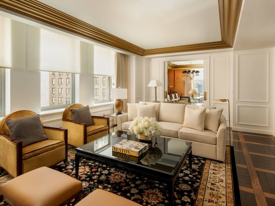A spacious living room with a beige sofa, two armchairs, a coffee table with books, and a large window with blinds.