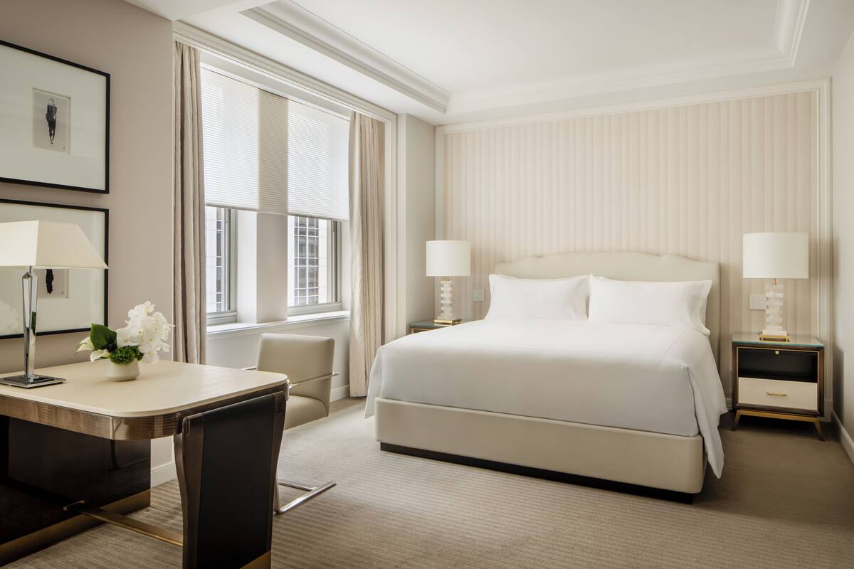 A hotel room with a large bed, two bedside tables with lamps, a desk with a chair, and a window with curtains.