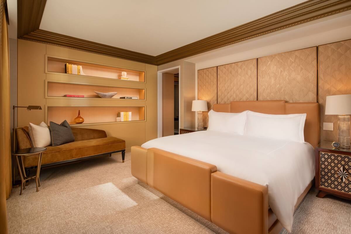A hotel room with a large bed, a sofa, a side table, and a wall-mounted shelf with books and decorative items.
