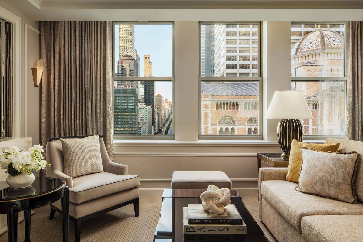A modern hotel room with a sofa, armchair, and a coffee table with books. Large windows offer views of city buildings and a historic structure.