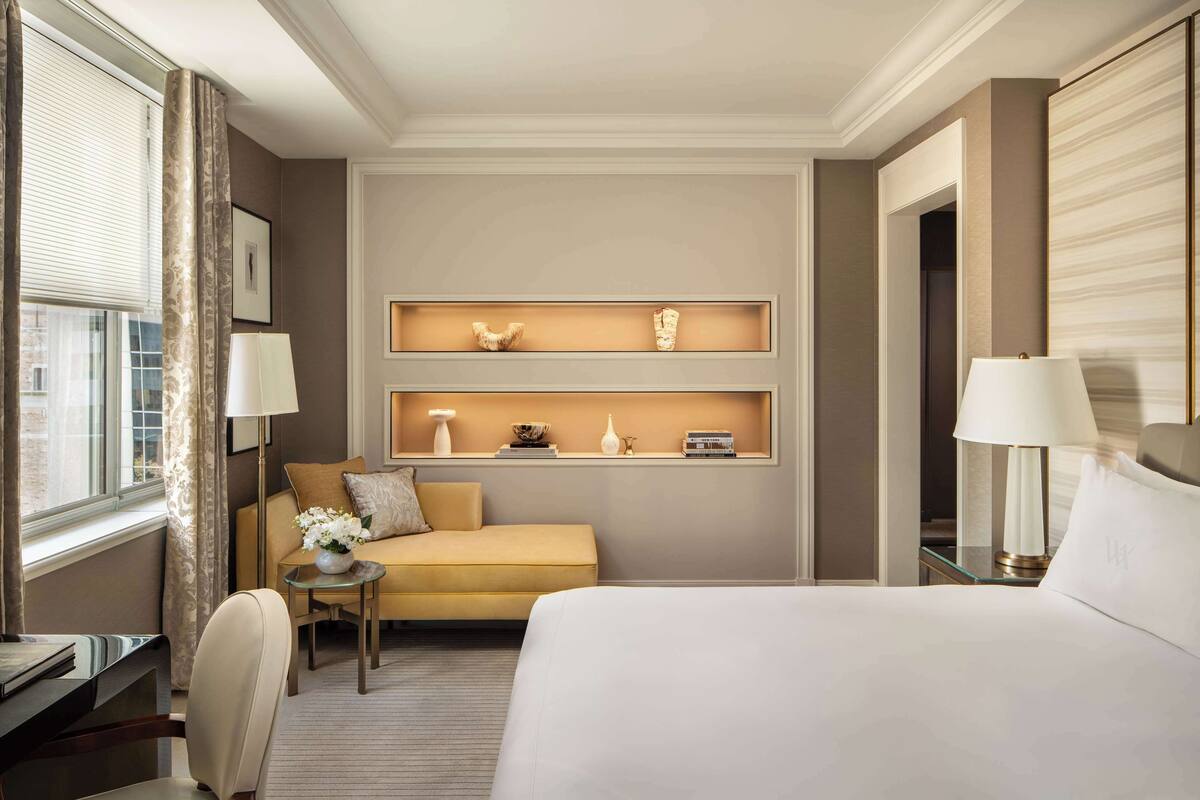 A hotel room with a bed, a sofa, a desk, and a small table with a lamp. There are wall-mounted shelves with decorative items.