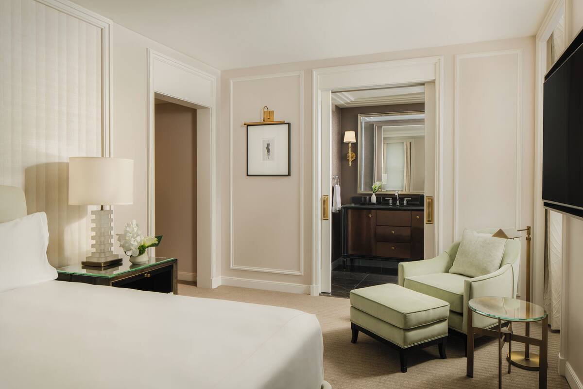 A hotel room with a bed, a nightstand with a lamp, a chair, a small table, and a bathroom visible through an open door.