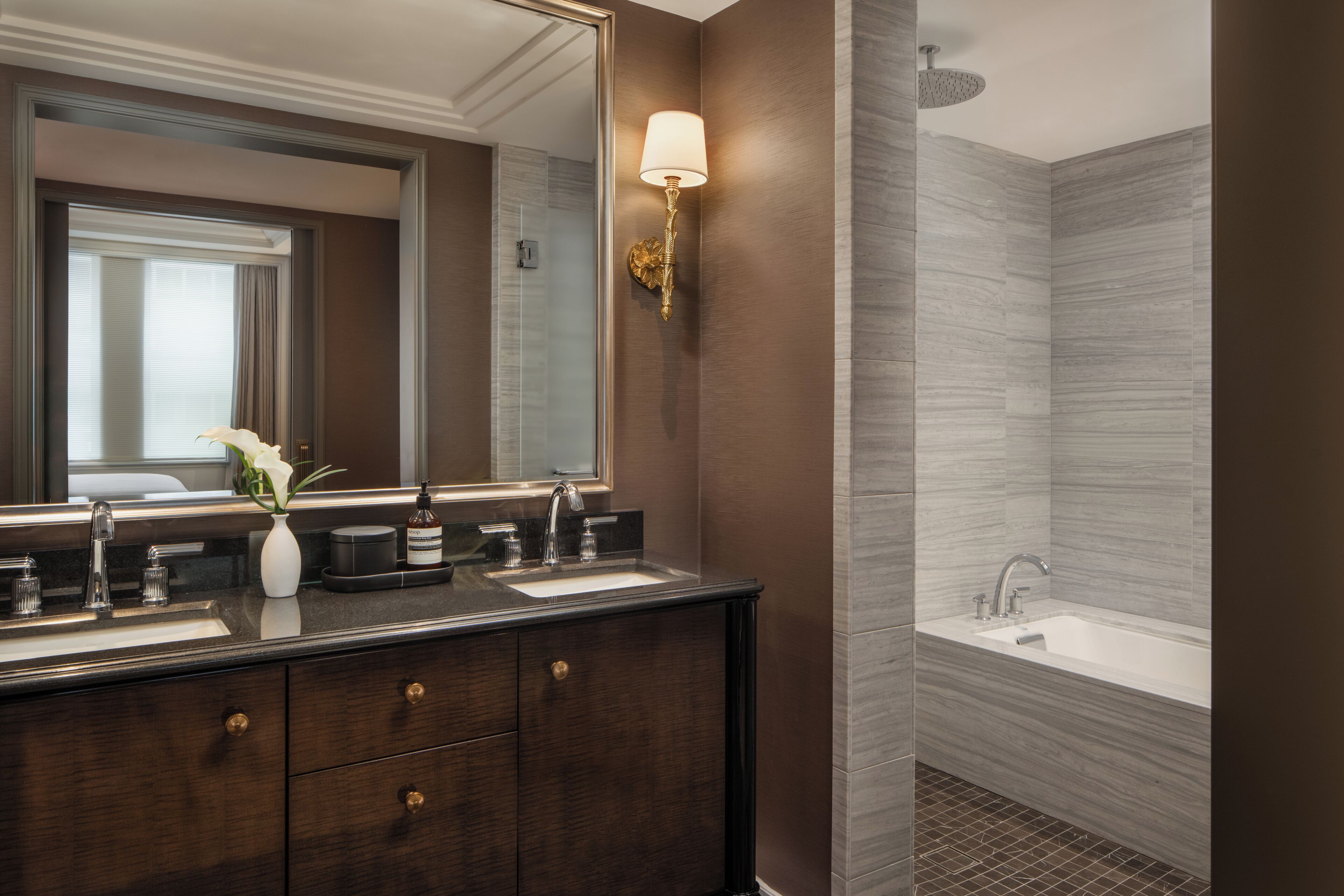 A bathroom with a dark wood vanity, a large mirror, a wall-mounted light, a bathtub, and a shower area.