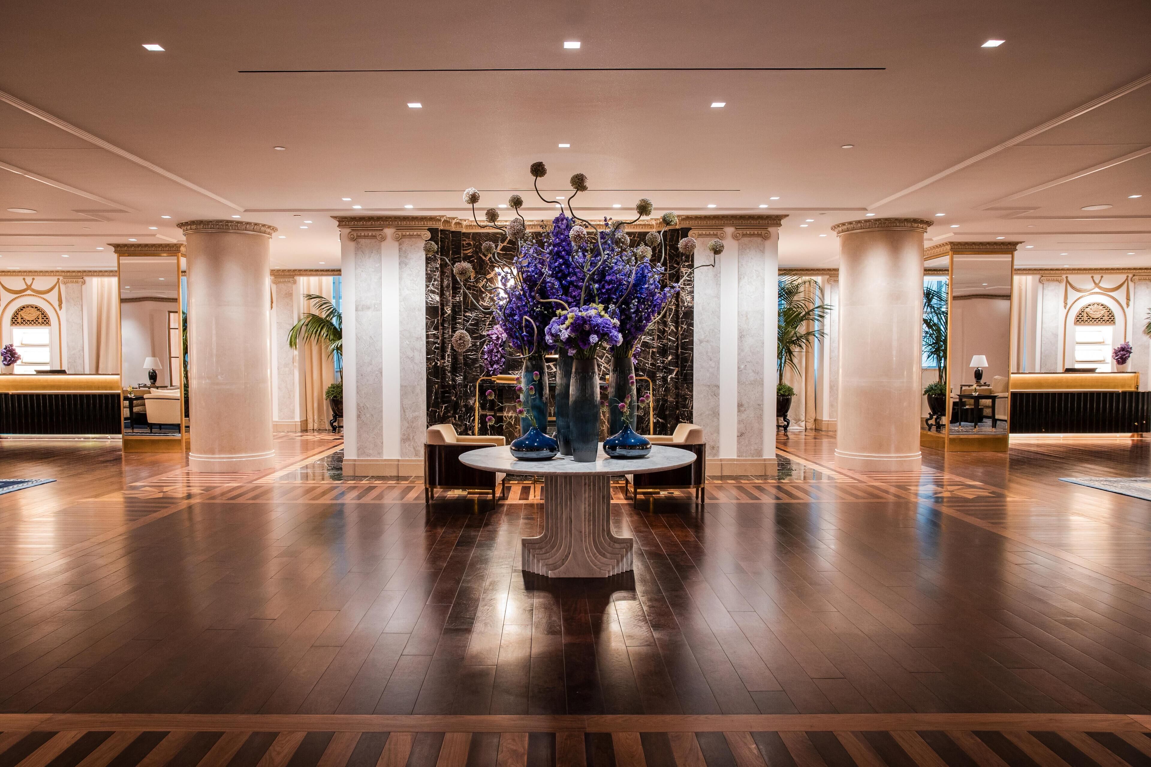 A spacious lobby with a central round table, flanked by two chairs, and a decorative floral arrangement.