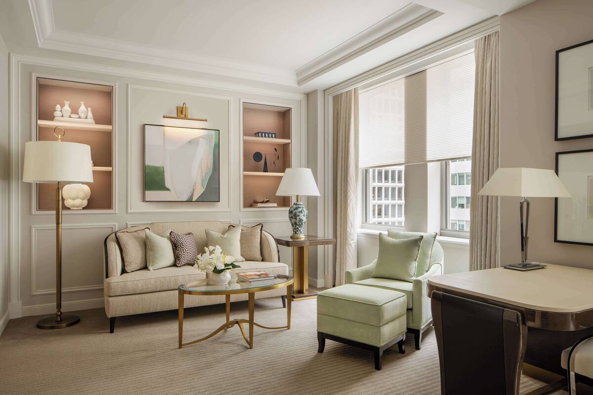 A well-lit living room with a sofa, armchairs, a coffee table, and a dining table. Shelves with decorative items and a painting are on the wall.