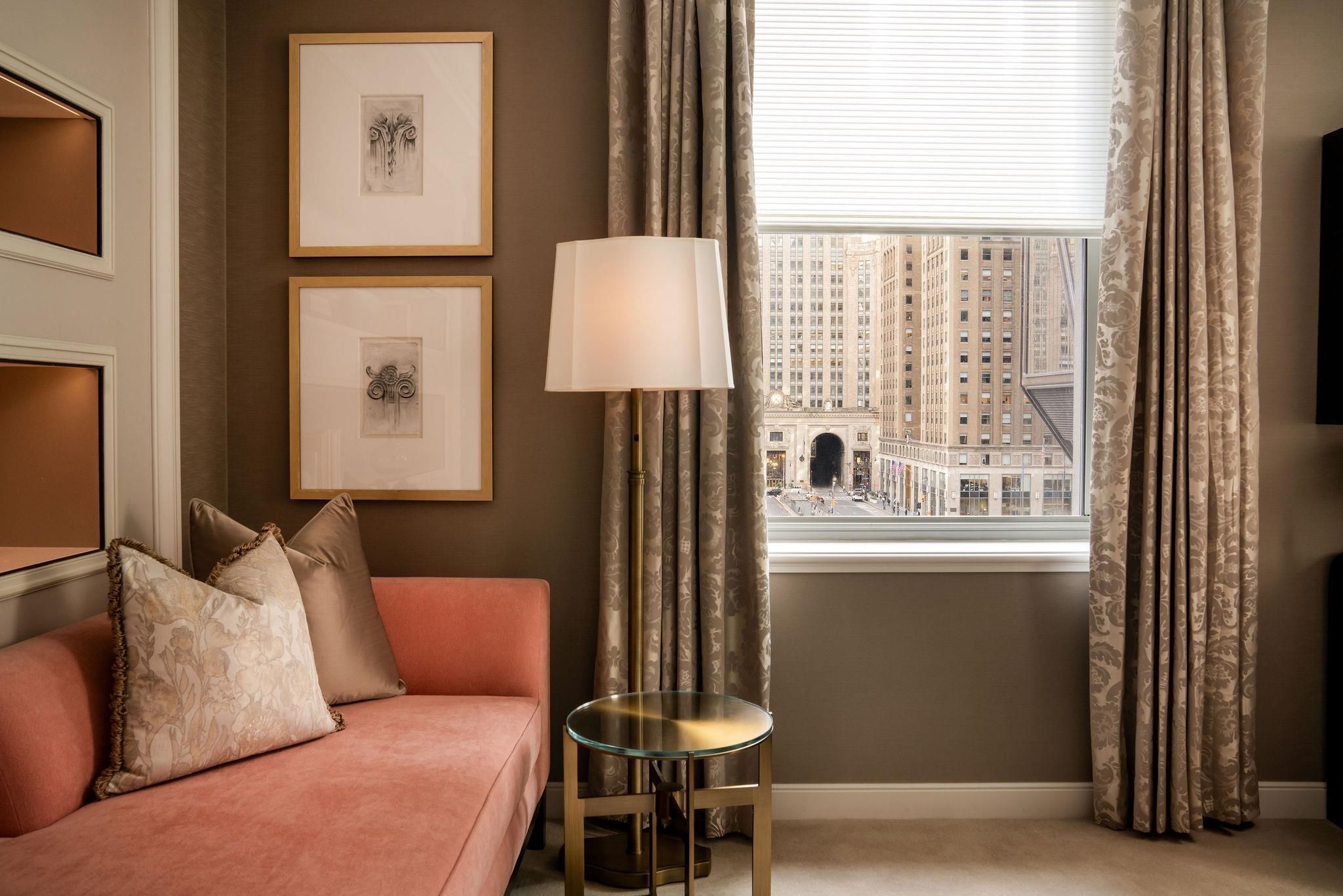 A room with a pink sofa, a glass table, framed artwork, curtains, and a view of a cityscape through a window.