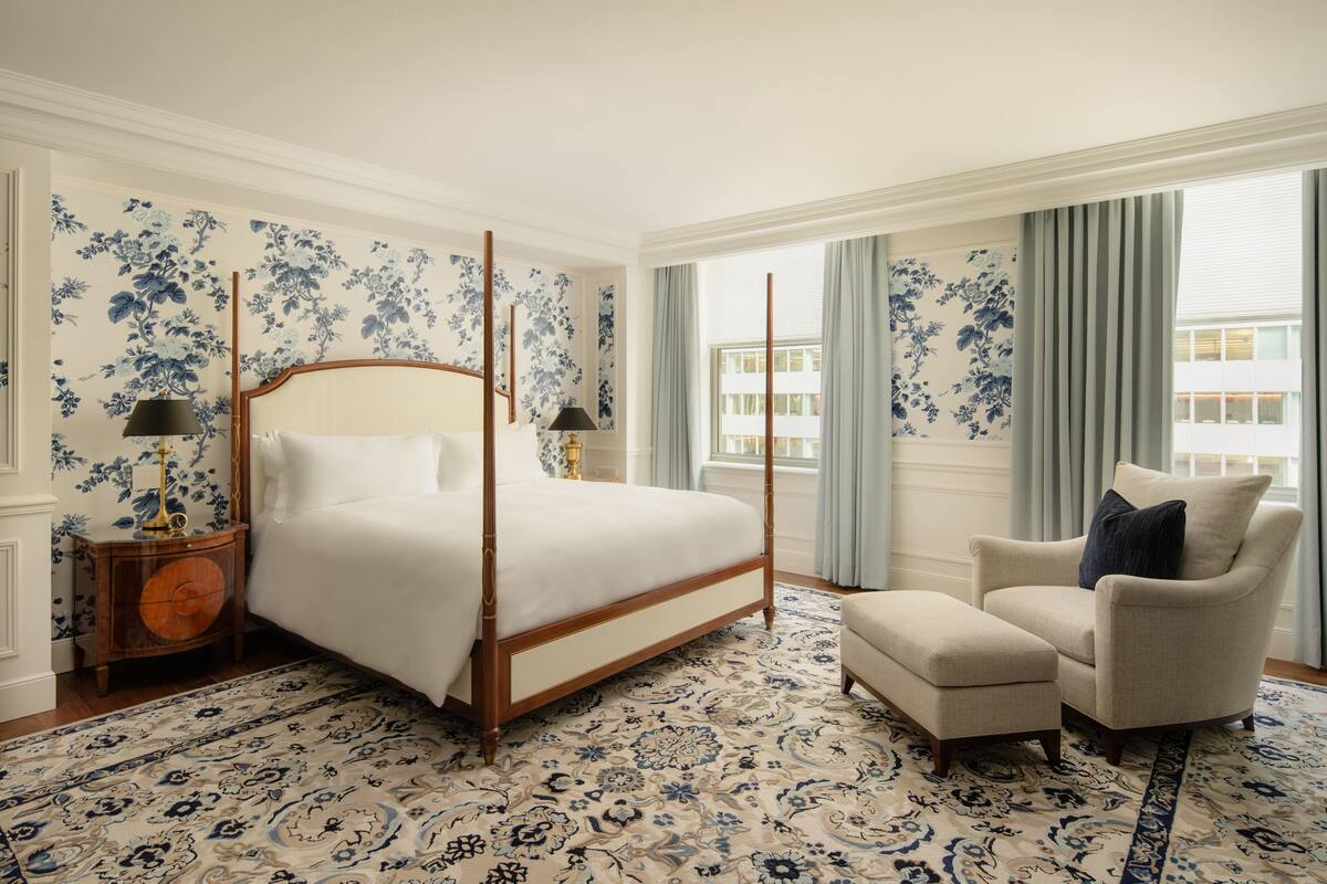 A four-poster bed with a wooden frame, a matching nightstand, a beige armchair, and a patterned rug.