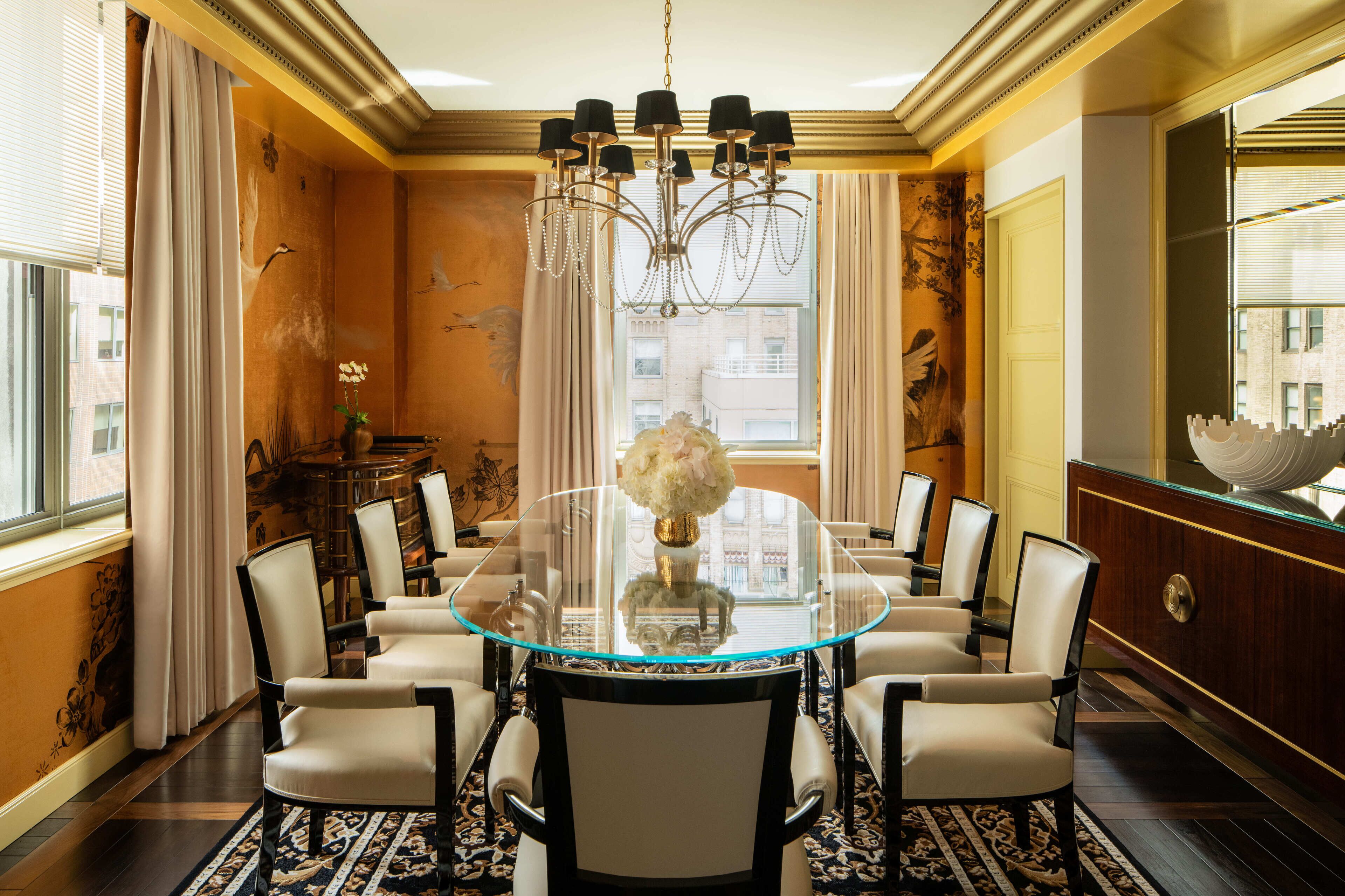 A dining area with a round glass table, six chairs, a chandelier, and a vase with flowers.