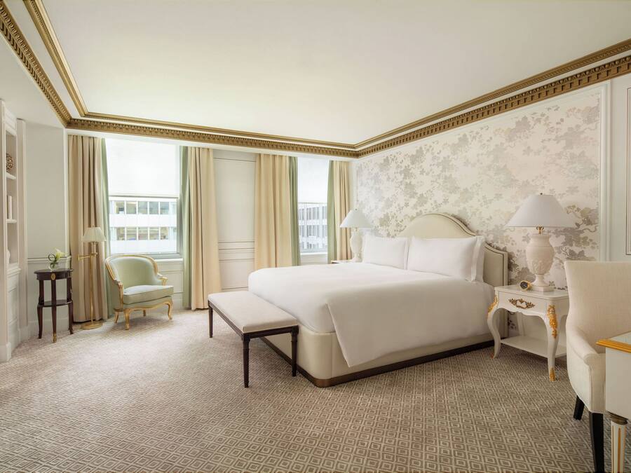 A hotel room with a large bed, a bedside table with a lamp, a chair, and a small table with a lamp.