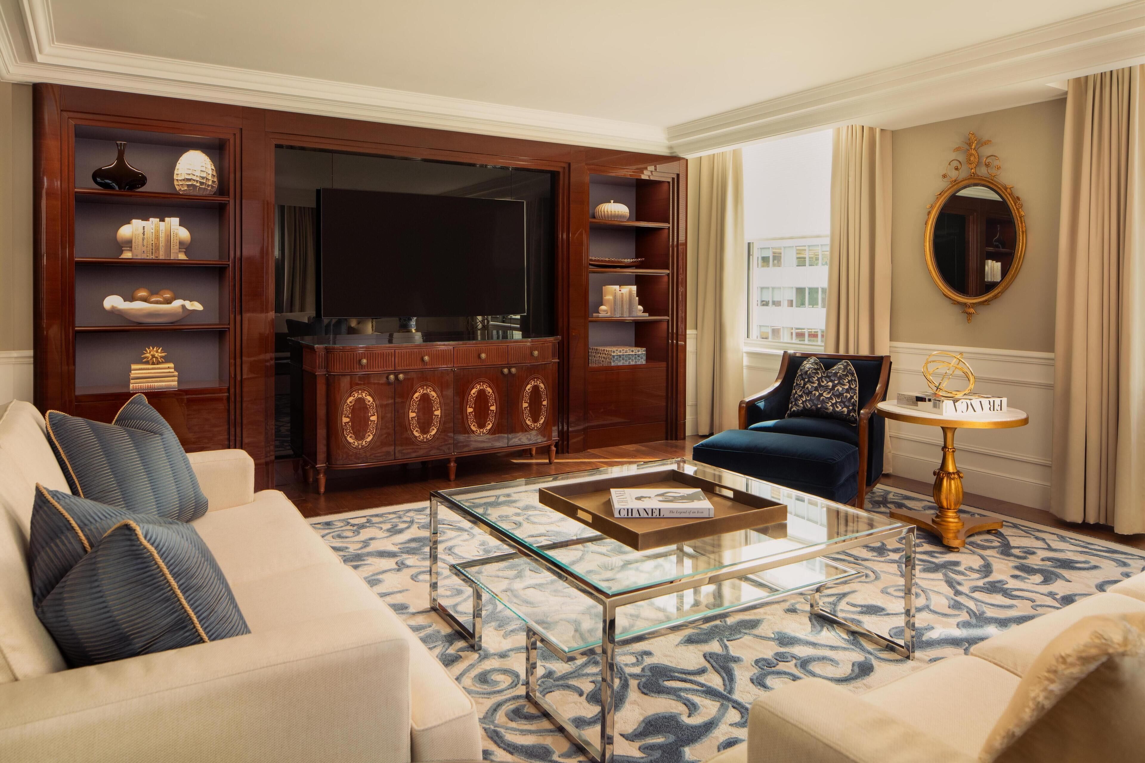 A spacious living room with a large flat-screen TV, a wooden entertainment unit, a blue sofa, and a glass coffee table.