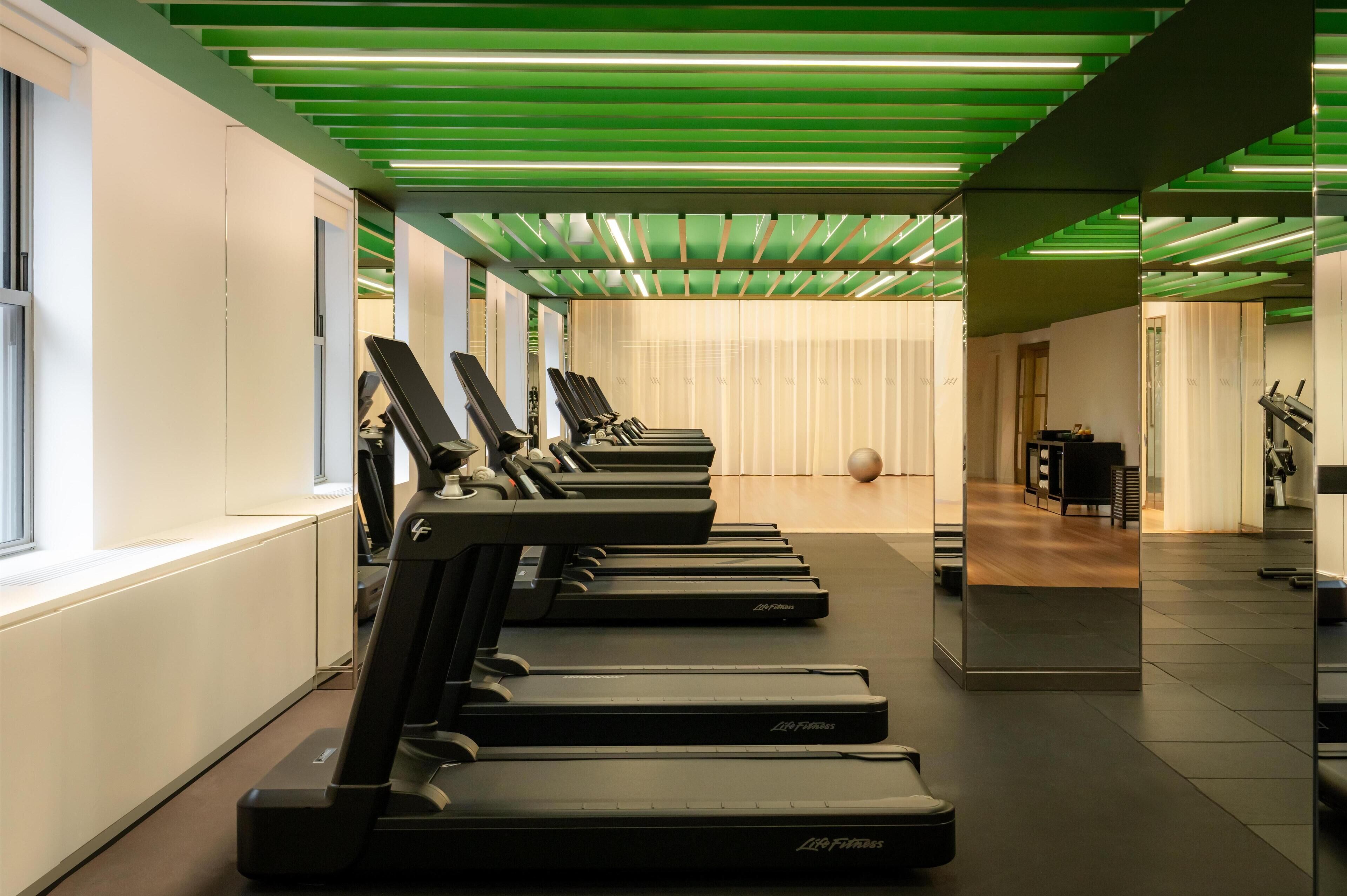 A gym with treadmills aligned along a wall, a mirrored wall reflecting the space, and a green-lit ceiling.
