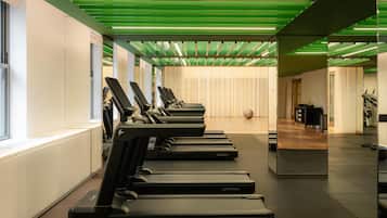 A gym with treadmills aligned along a wall, a mirrored wall reflecting the space, and a green-lit ceiling.