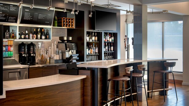 A modern bar area with a wooden bar counter, multiple stools, and a variety of liquor bottles. - Courtyard New Haven Wallingford (Wallingford)
