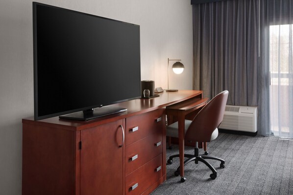 Habitación de hotel con escritorio de madera, televisor de pantalla plana, lámpara, silla y ventana grande con cortinas.