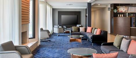 A modern hotel lobby with a large flat-screen TV, comfortable seating, a Starbucks coffee station, and a patterned carpet.