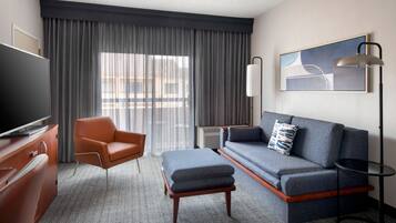 Ein Hotelzimmer mit Sofa, Hocker, Sessel, Fernseher und Kunstwerk an der Wand.