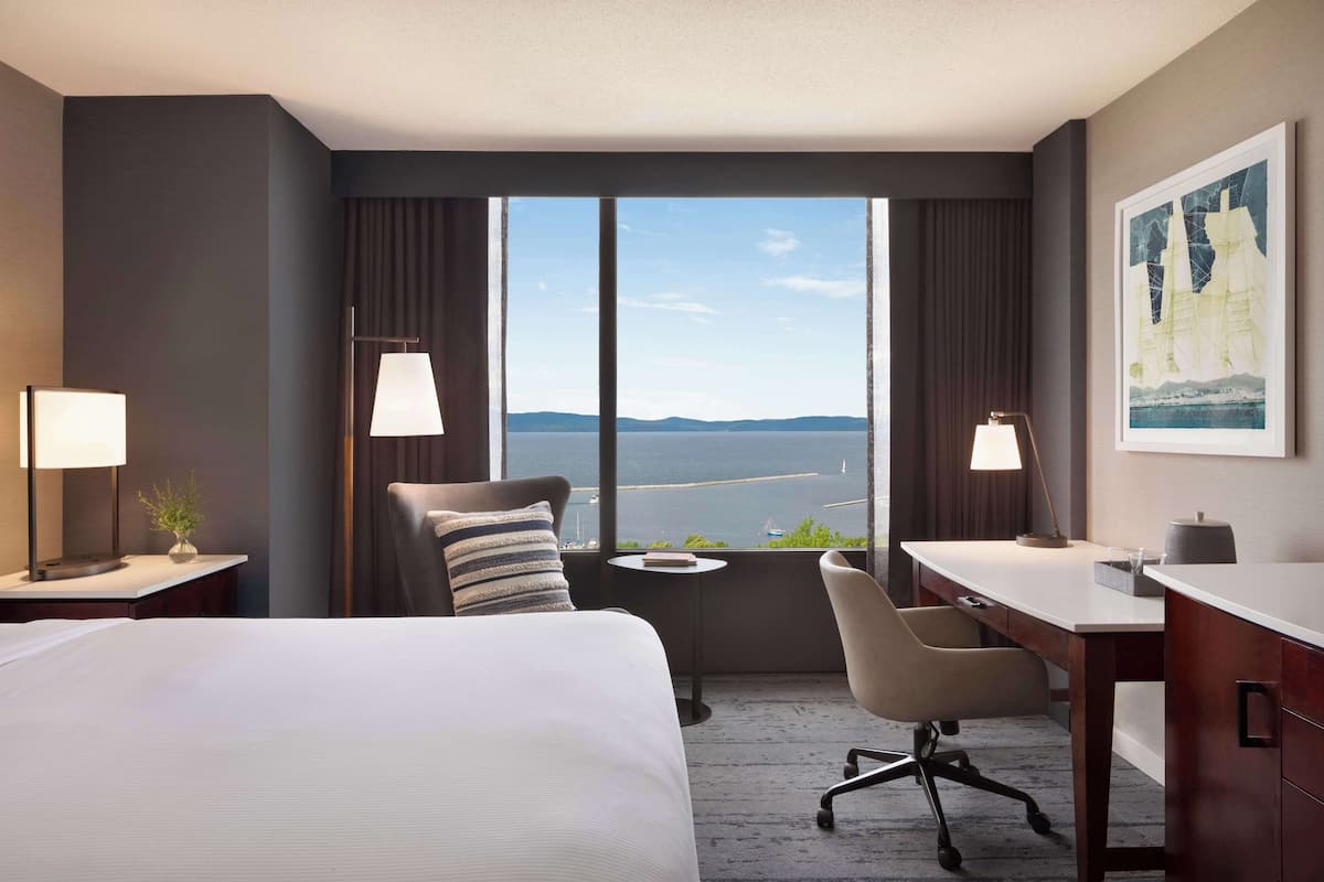 A hotel room with a bed, a desk, a chair, and a view of the sea through large windows.