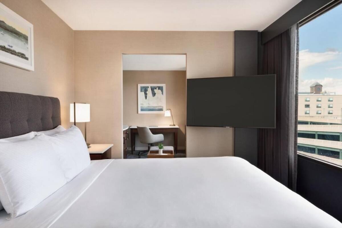 A hotel room with a bed, a desk, a chair, a TV, and a window with a city view.