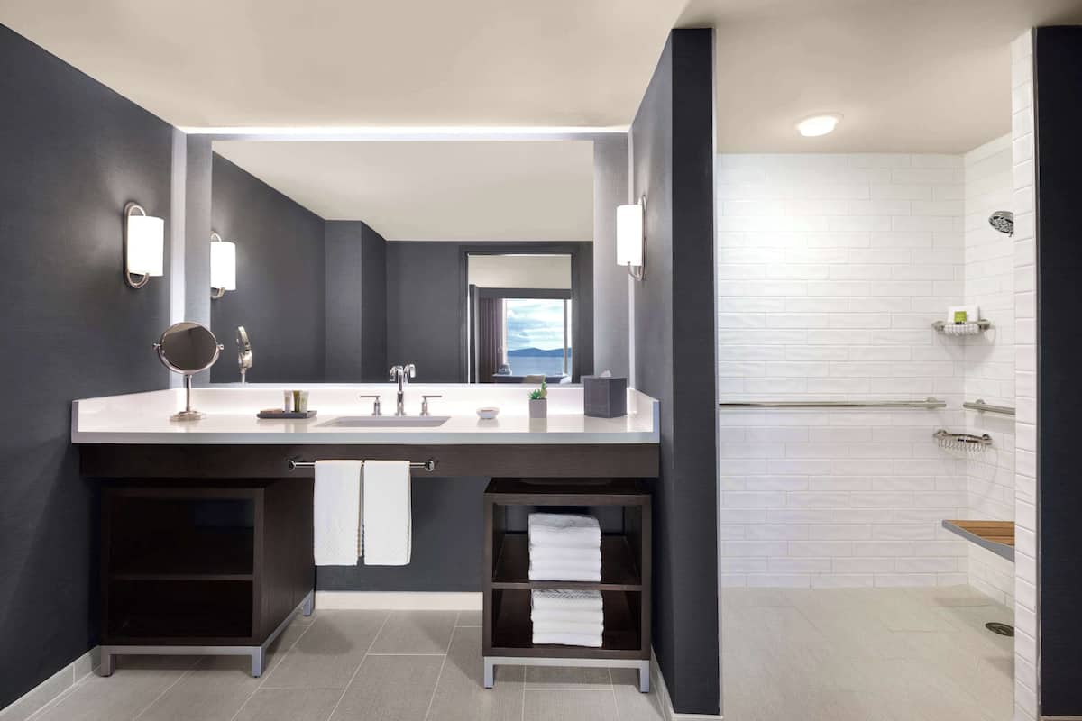 A modern bathroom with a dark gray and white color scheme, featuring a large mirror, dual sinks, and a shower area.