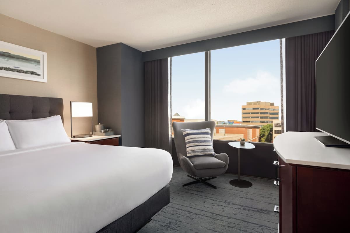 A hotel room with a large bed, a chair, a desk, a TV, and a view of the city.