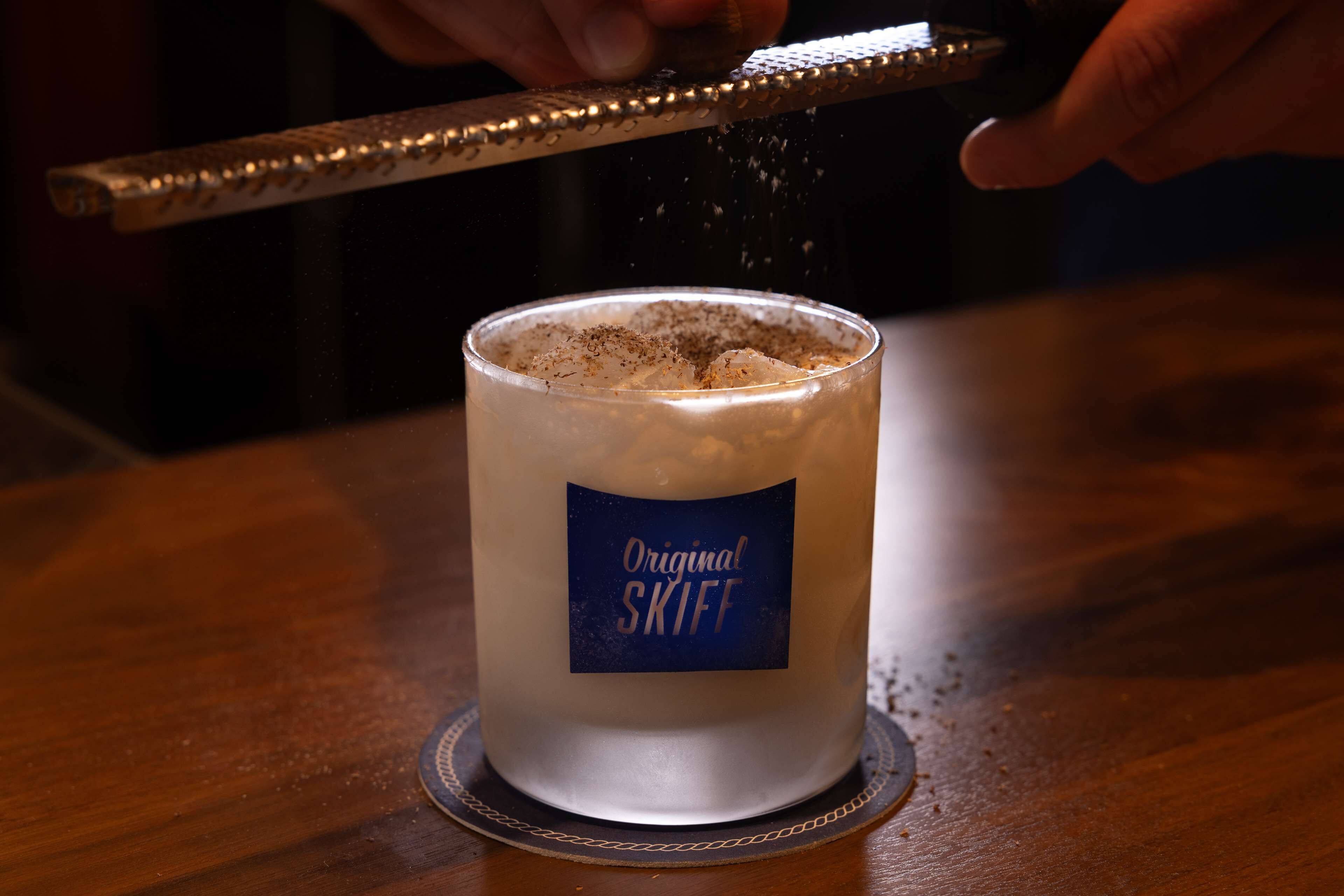 A hand grating a substance over a glass labeled "Original Skiff."