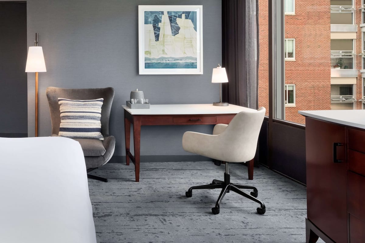A hotel room with a bed, a desk, a chair, a lamp, a picture on the wall, and a view of a brick building.
