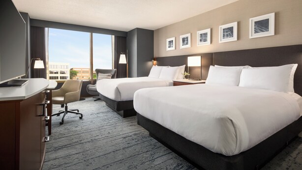 Habitación de hotel con dos camas, un escritorio, una silla y un ventanal con vistas a la ciudad.