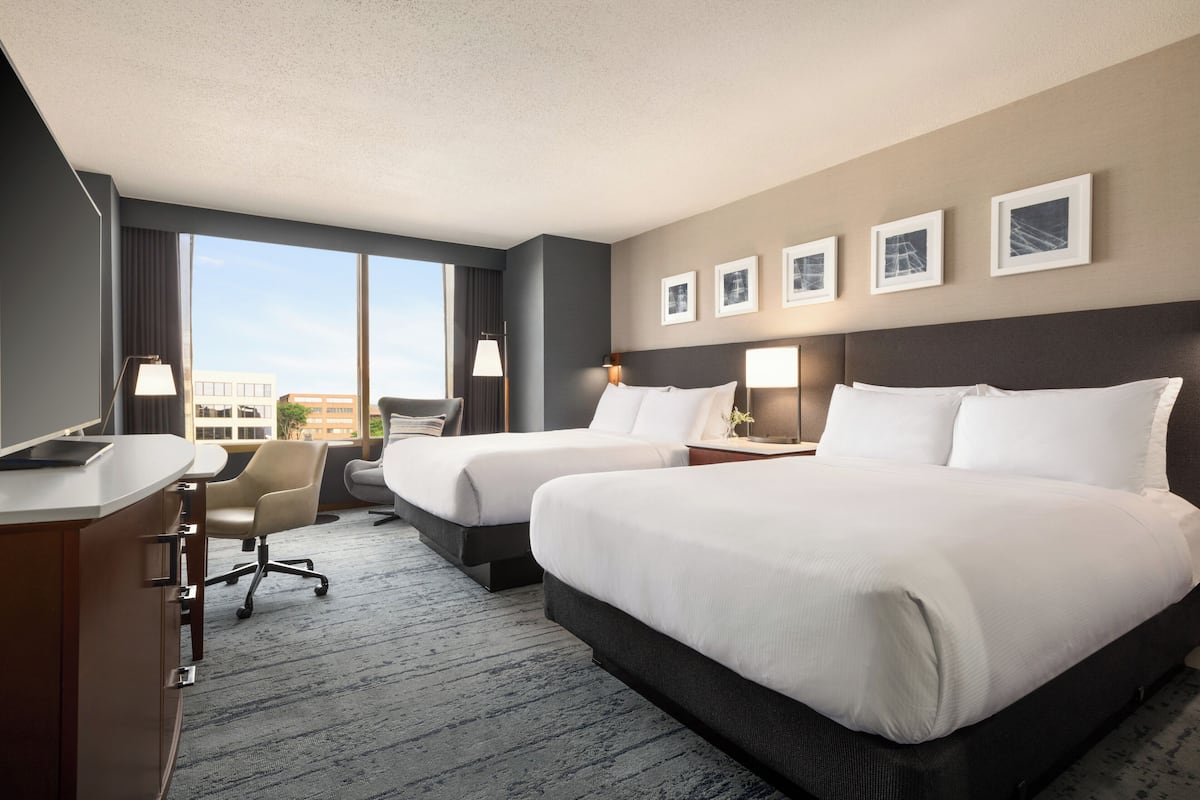 A hotel room with two beds, a desk, a chair, and a large window offering a city view.